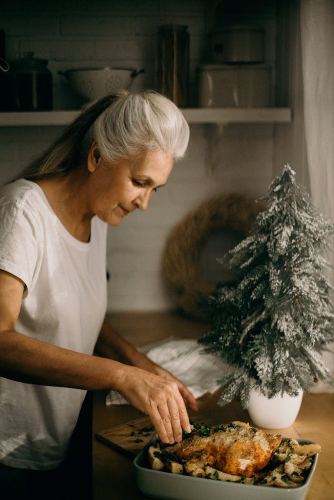 Senior woman garnishing a roast dish beside a small snowy tree for holiday dinner.